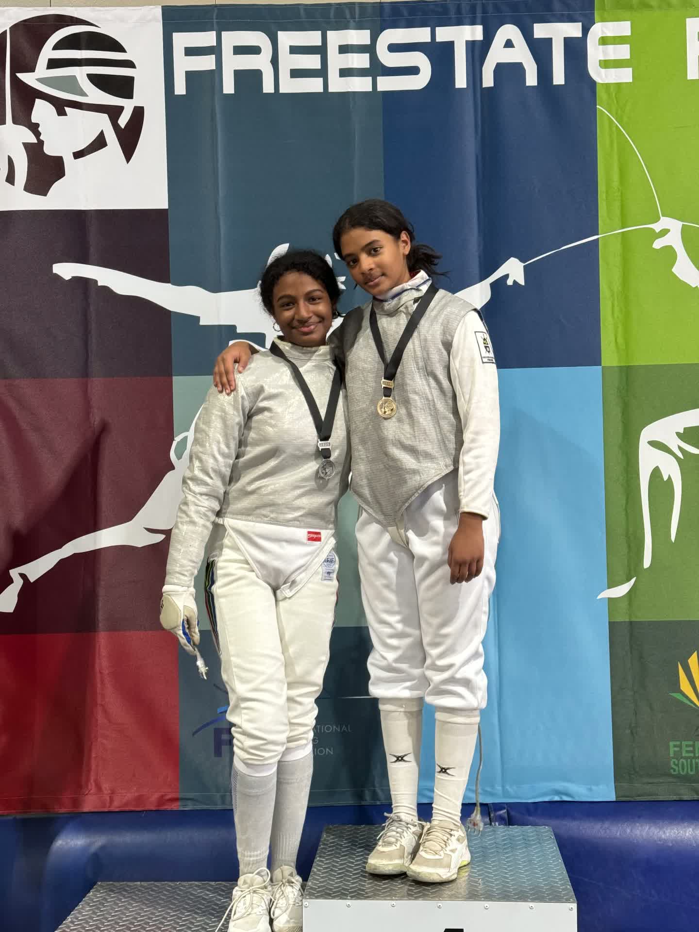Danica and Gianna McIntosh with their Free State Open medals in front of the Freestate Fencing banner